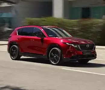 Front three quarter view of a Soul Red Mazda CX-5 SUV driving on a city street past an office building