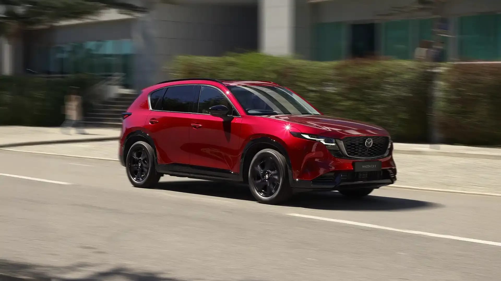 Front three quarter view of a Soul Red Mazda CX-5 SUV driving on a city street past an office building