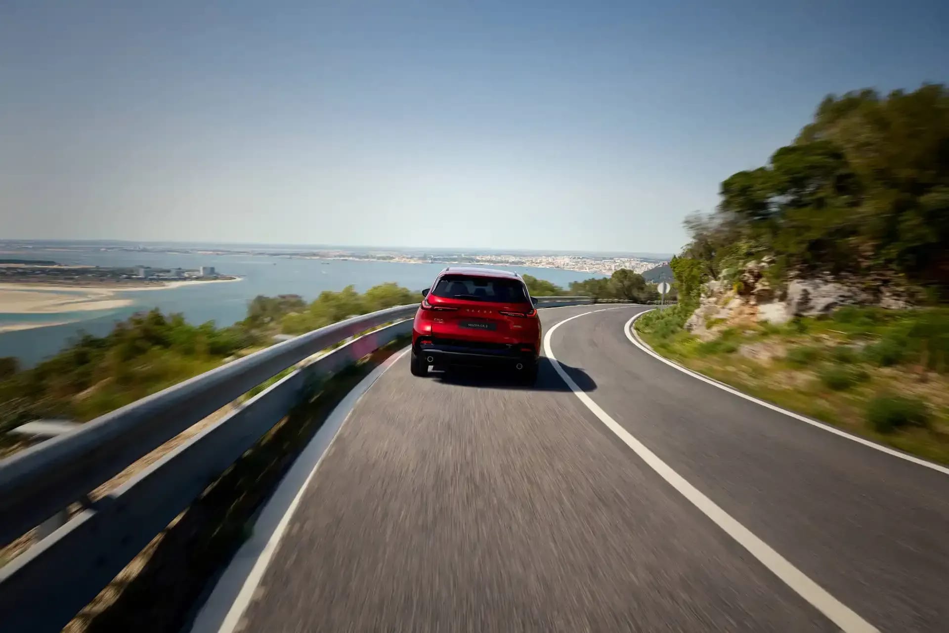 Rear view of a red Mazda CX-5 SUV driving on an open road near a harbour