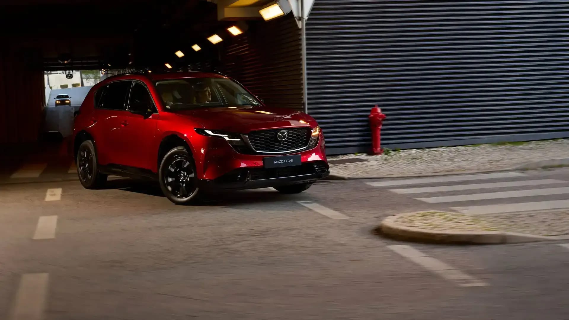 Front three quarter view of a Soul Red Mazda CX-5 SUV driving around a corner exiting a tunnel in a city street