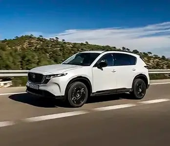Front three quarter view of a white all-new Mazda CX-5 SUV driving on an open road with a tree-covered hill and blue skies in the background