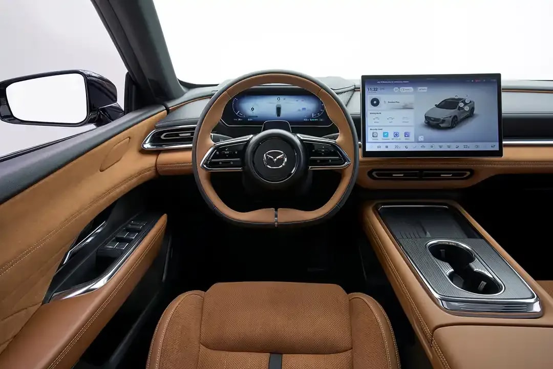 Interior view of the tan alcantara front seat, steering wheel and dashboard - featuring a large centre touchscreen - of the Mazda 6e electric car