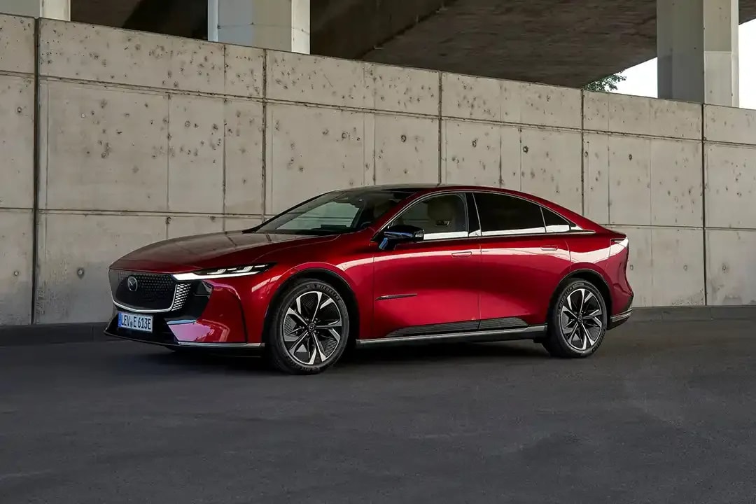 Front side view of a Soul Red Mazda 6e electric car parked next to a concrete wall under a freeway overpass