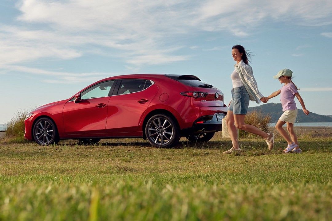 A parent and child walking past their Soul Crystal Red Metallic Mazda3 on the way to a picnic on the beach