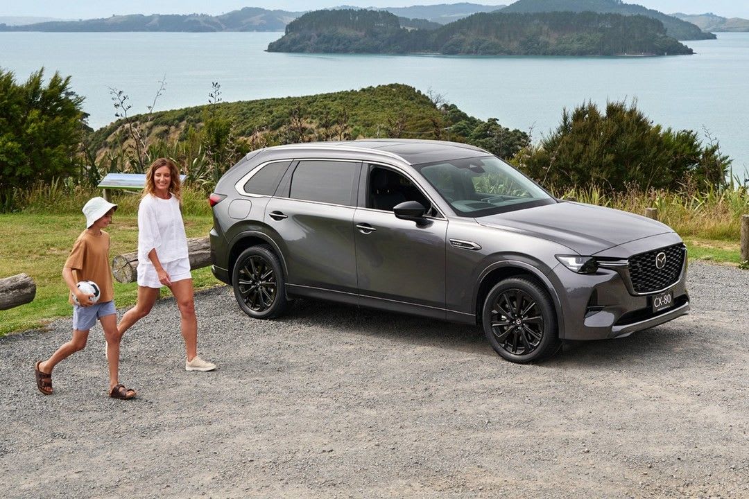 A parent and child walking towards their Machine Grey Metallic Mazda CX-80 in a gravel parking lot overlooking the water