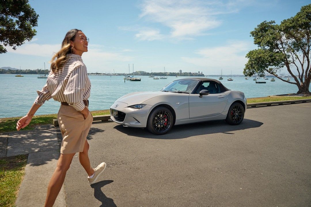 A person strolling towards their Aero Grey Metallic Mazda MX-5 Roadster, parked by the waterside