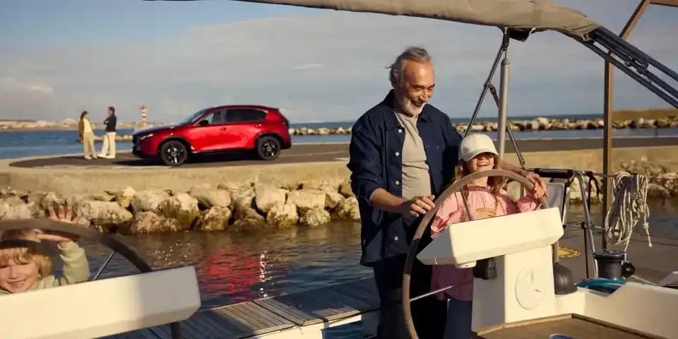 Man and girl at the wheel of a yacht in a marina with their Soul Red Mazda CX-5 SUV parked in the background