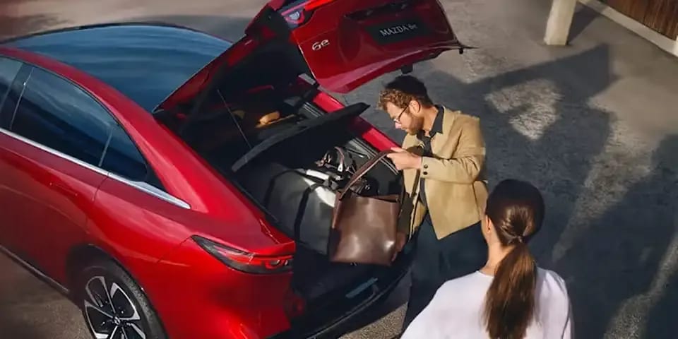 Top rear view of a man loading luggage into the boot of a Soul Red all-electric Mazda 6e hatchback