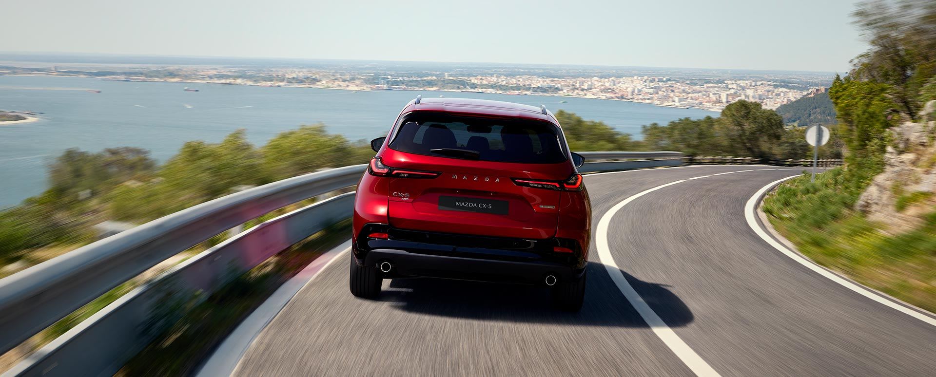 Rear view of a Soul Red Mazda CX-5 driving on a winding mountain road with a harbour and city visible below