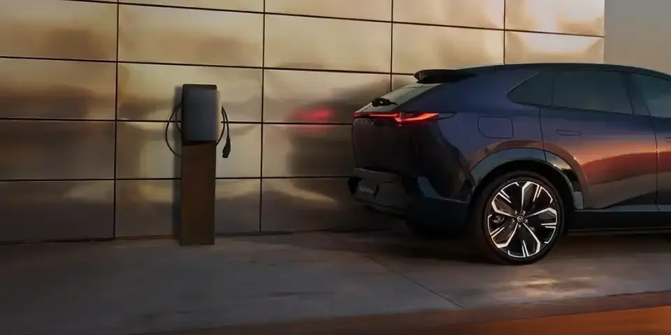 Rear half of a Nightfall Violet all-electric Mazda CX-6e SUV shown parked next to a charge point in a driveway