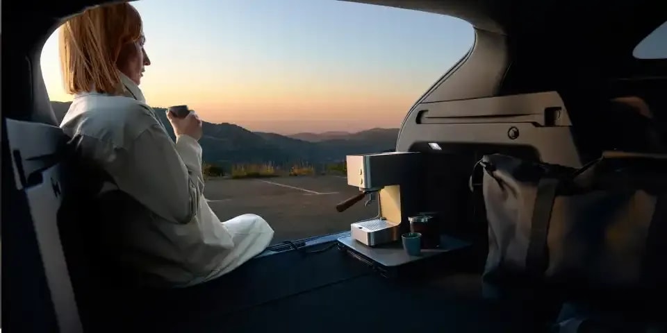 Looking out of the boot of an all-electric Mazda CX-6e with a woman sitting in the boot next to her portable coffee machine plugged into the car