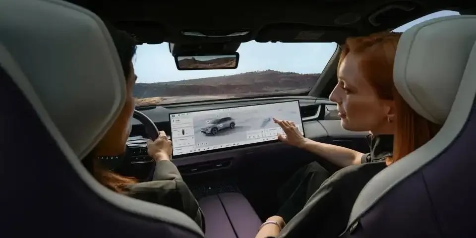 Man and woman in the front seats of the all-electric Mazda CX-6e SUV with the woman gesturing to the touchscreen for easy control of the vehicle functions