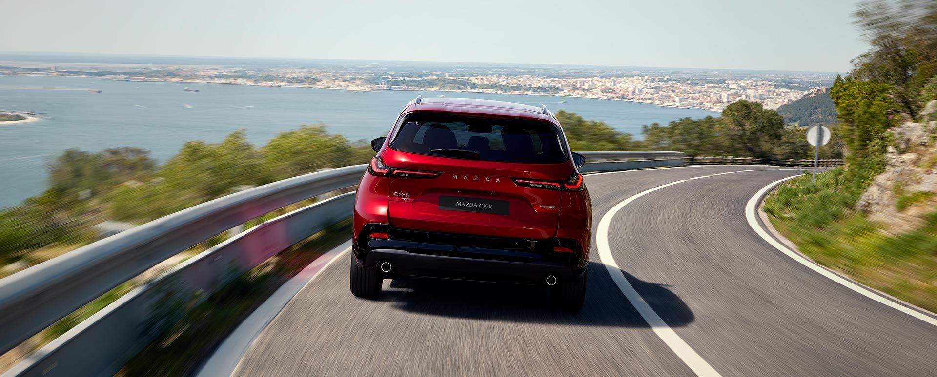 Rear view of a Soul Red Mazda CX-5 driving on a winding mountain road with a harbour and city visible below