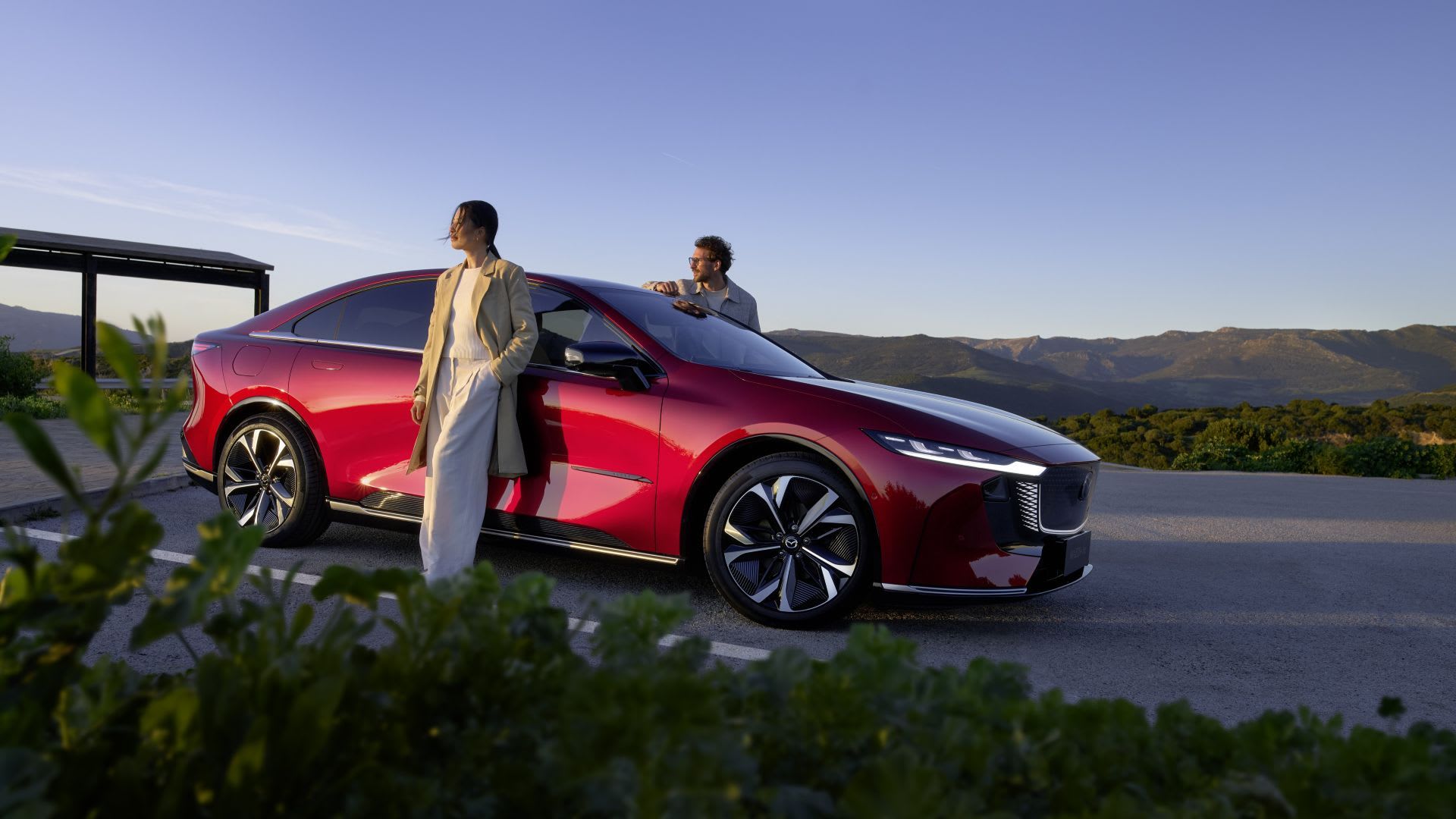 Front three quarter view of a Soul Red all electric Mazda 6e hatchback with a woman and man standing alongside the car