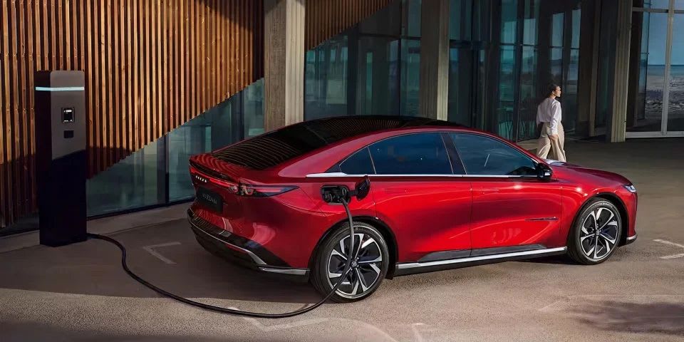 Rear three quarter view of a Soul Red all-electric Mazda 6e hatchback recharging outside an office building for driver convenience