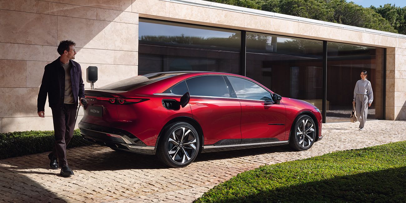Rear three quarter view of a Soul Red all-electric Mazda 6e hatchback with a man conveniently recharging it in his driveway