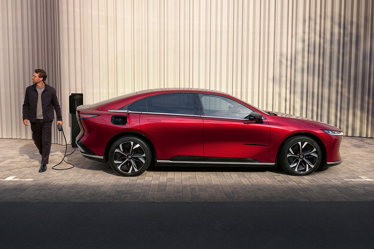 Side view of a Soul Red all-electric Mazda 6e hatchback being recharged by a man at a public charging station