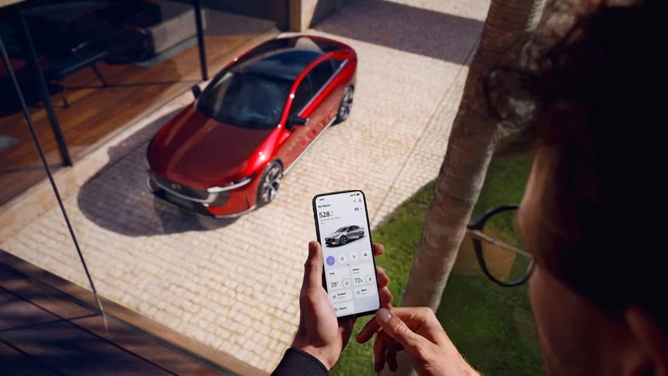 Man looking at the Mazda App on his smartphone with his Soul Red all-electric Mazda 6e hatchback visible in the background 