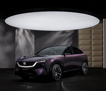 Front three quarter view of the all new, all-electric Mazda CX-6e SUV in a deep purple colour on display under a large spotlight