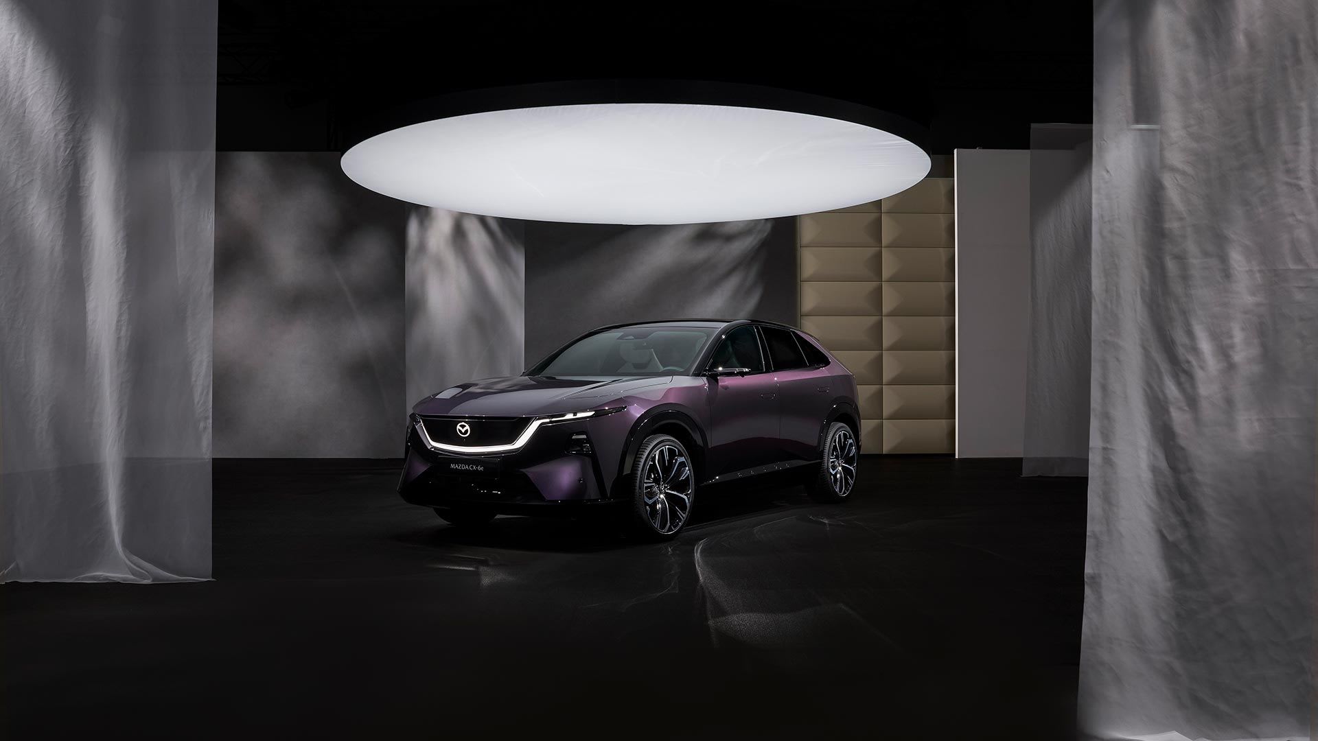 Front three quarter view of the all new, all-electric Mazda CX-6e SUV in a deep purple colour on display under a large spotlight
