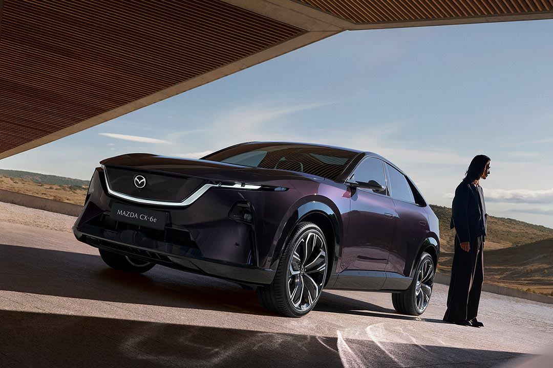 Front three quarter view of a deep purple all-electric Mazda CX-6e SUV with a woman standing next to the car