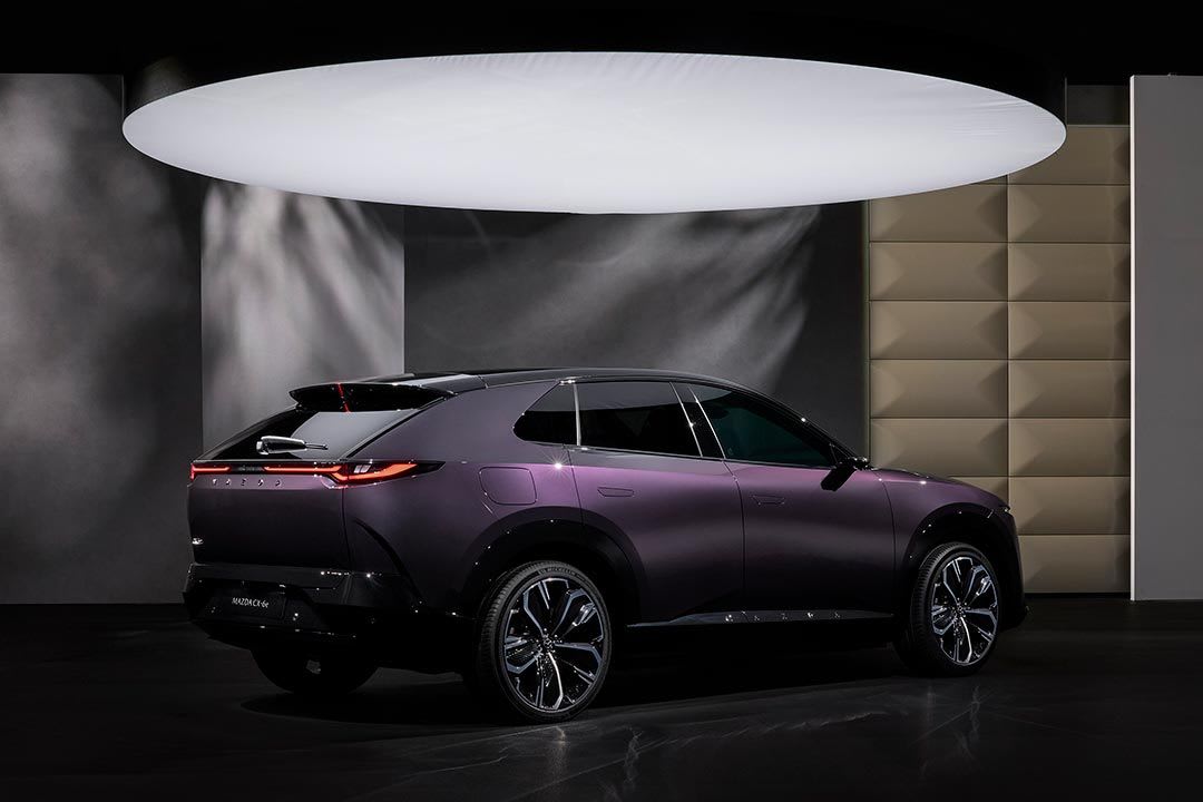 Rear three quarter view of a deep purple all-electric Mazda CX-6e SUV on display under a large spotlight
