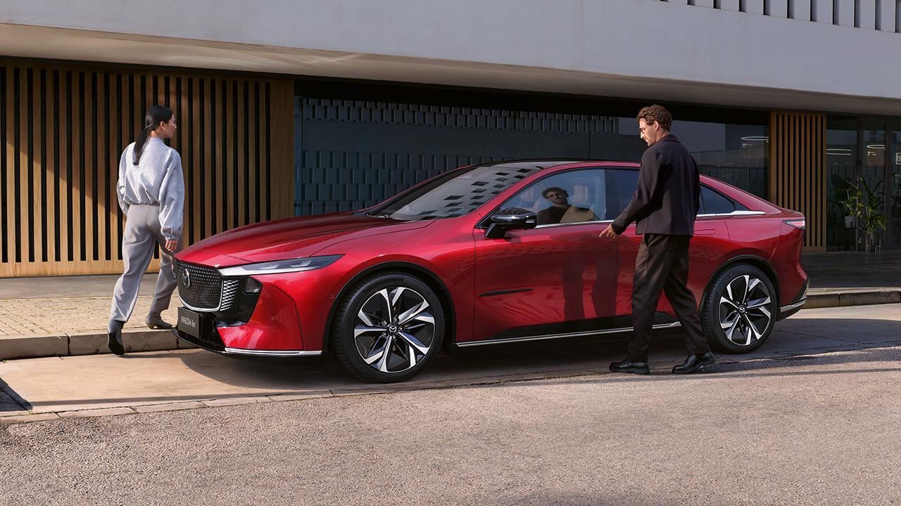 Front three quarter view of a Soul Red all-electric Mazda 6e hatchback parked in front of an office building with a man and woman entering the car