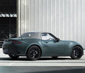 Rear three quarter view of a Zinc Green Mazda MX-5 Roadster parked in front of a large warehouse
