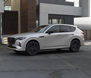 Front three quarter view of a Rhodium White Mazda CX-60 SUV hybrid parked in front of a designer home