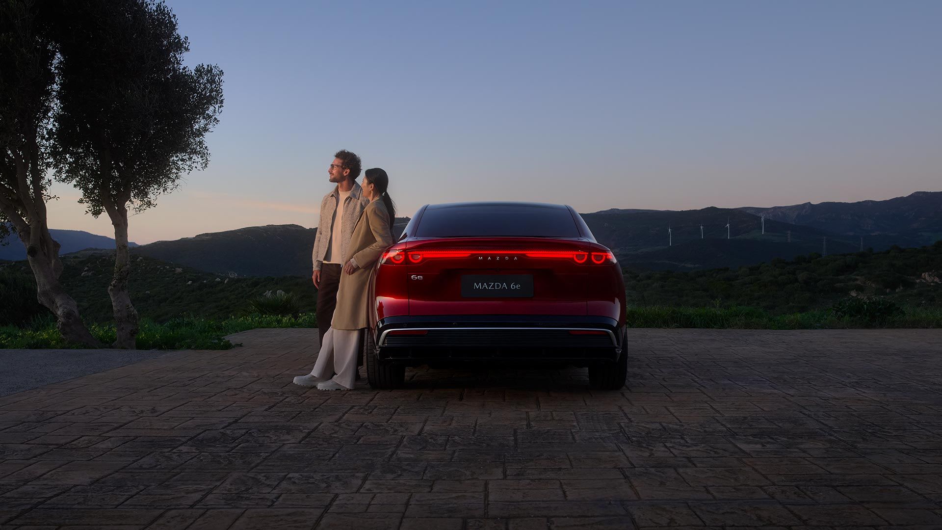 Rear view of a Soul Red all-electric Mazda 6e hatchback parked at dusk with a man and woman leaning against the car