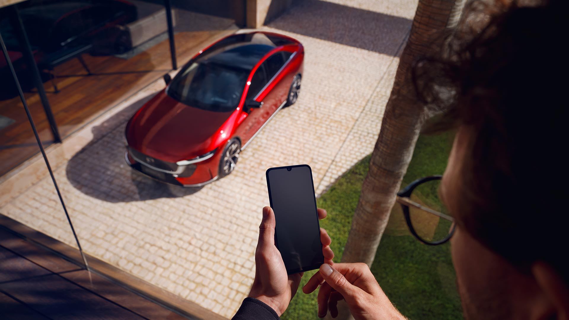 Man looking at his smartphone screen with a Soul Red all-electric Mazda 6e parked below in his driveway