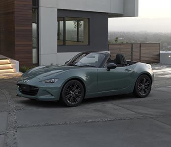 Front three quarter view of a Zinc Green Mazda MX-5 Roadster parked in front of an apartment building