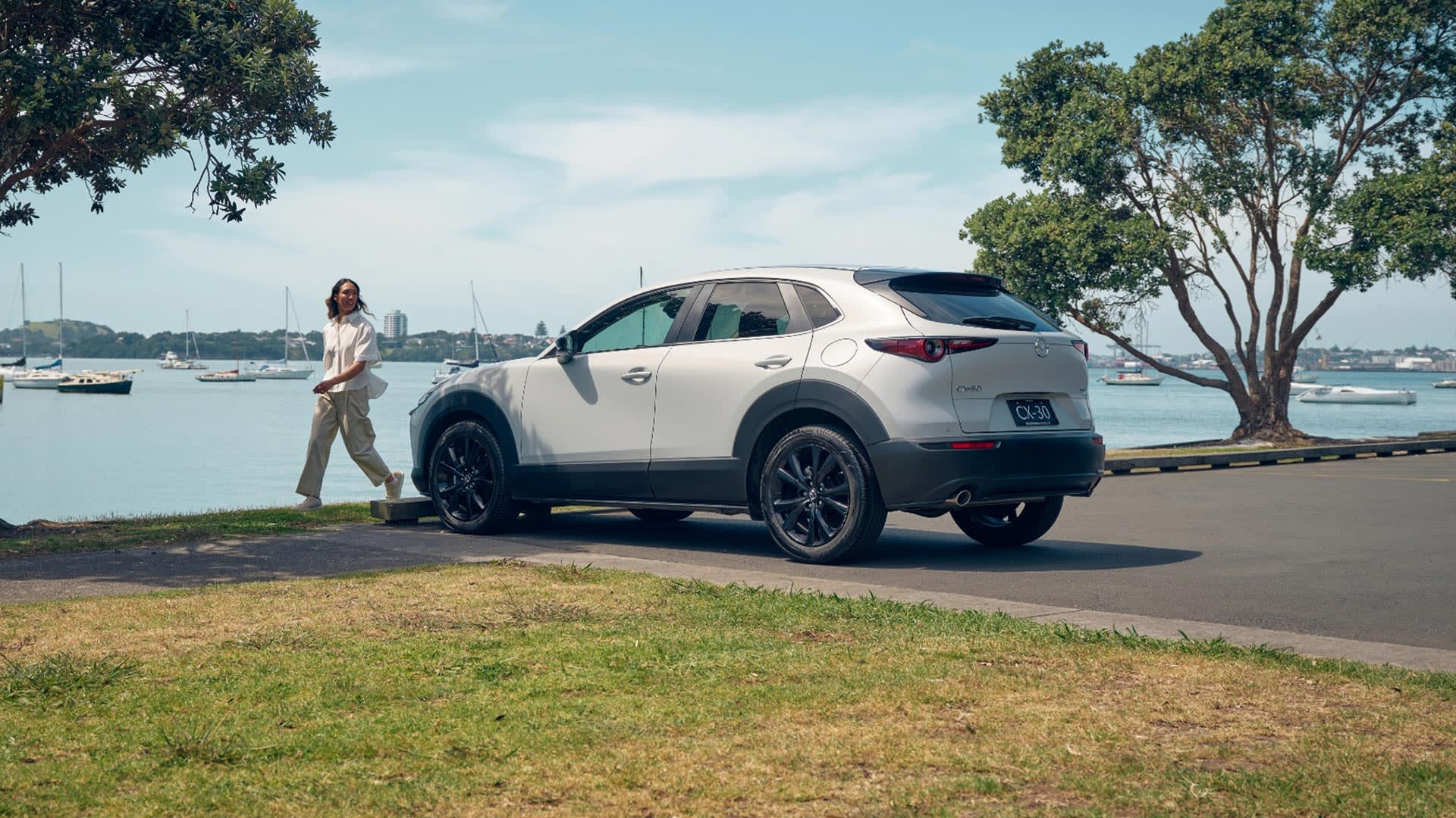 Rear three quarter view of a Snowflake White Mazda CX-30 SUV parked at a harbour with a woman walking by