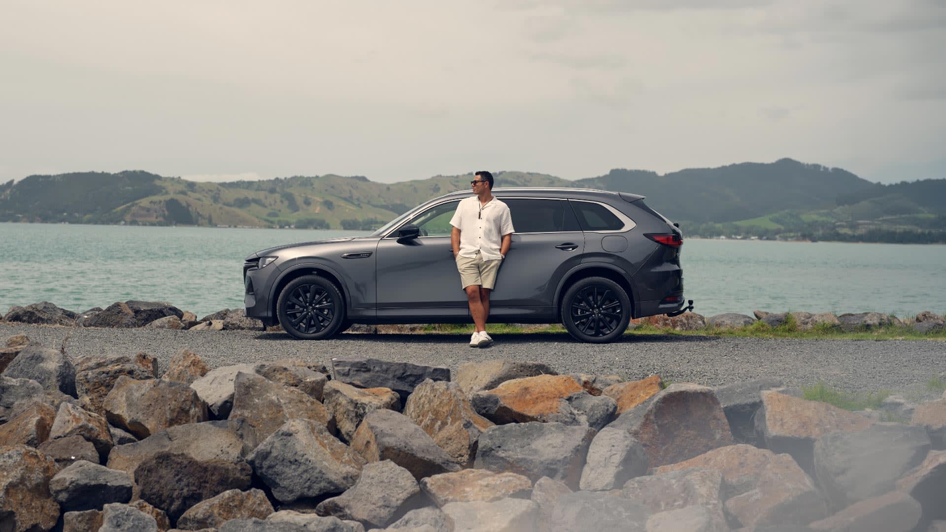 Side view of a Machine Grey Mazda CX-80 SUV parked on a breakwater with a man leaning against the car
