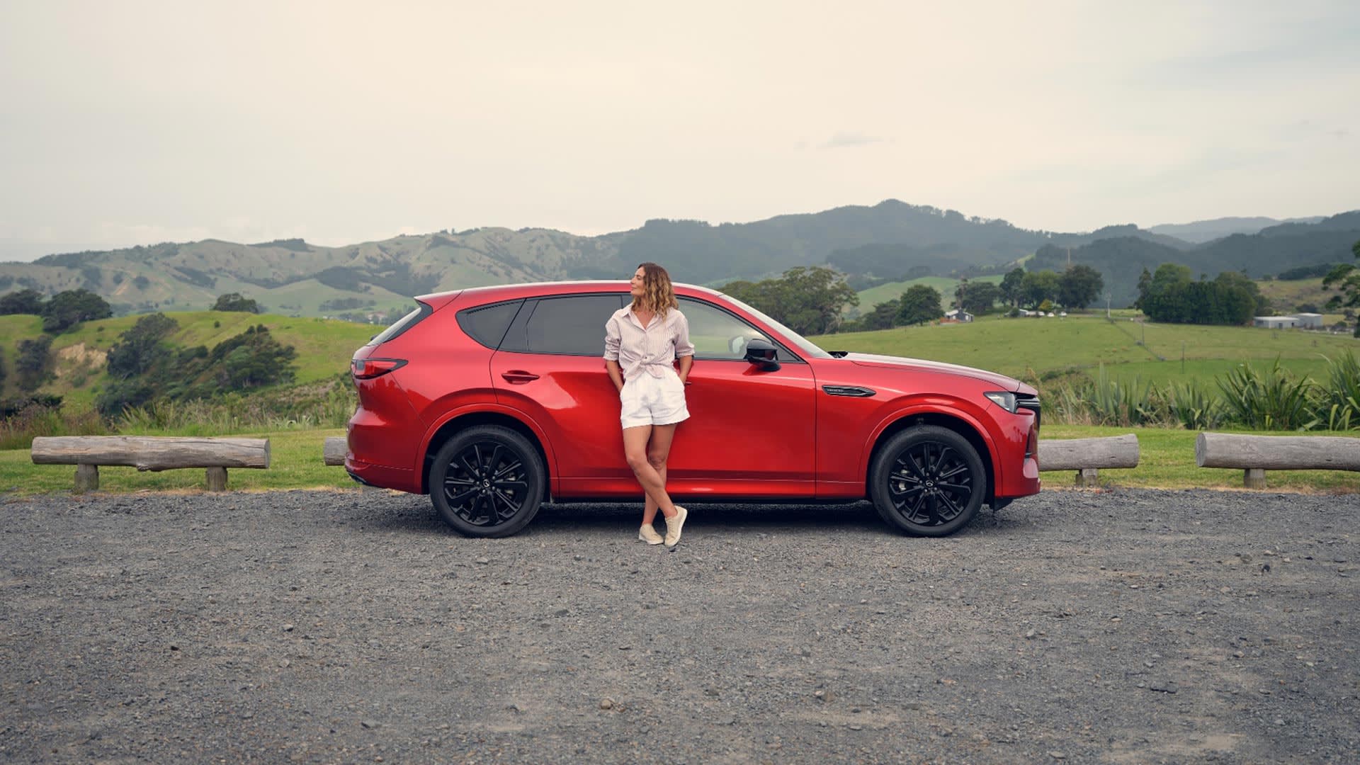 Side view of a Soul Red Mazda CX-60 SUV parked near green rolling hills with a woman leaning against the car