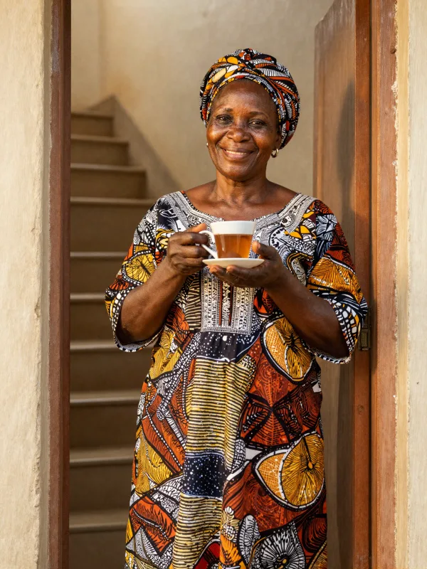A joyful Nigerian woman in her 50s holding a cup of ginger tea in her sunlit doorway