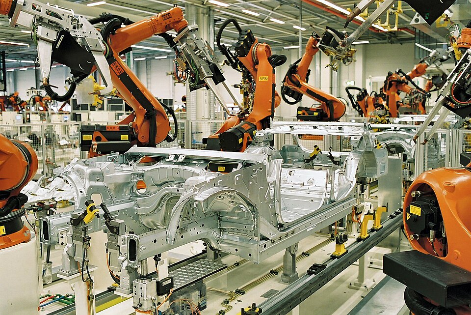 Rows of orange industrial robot arms working around a bare steel car body shell on an automobile assembly line