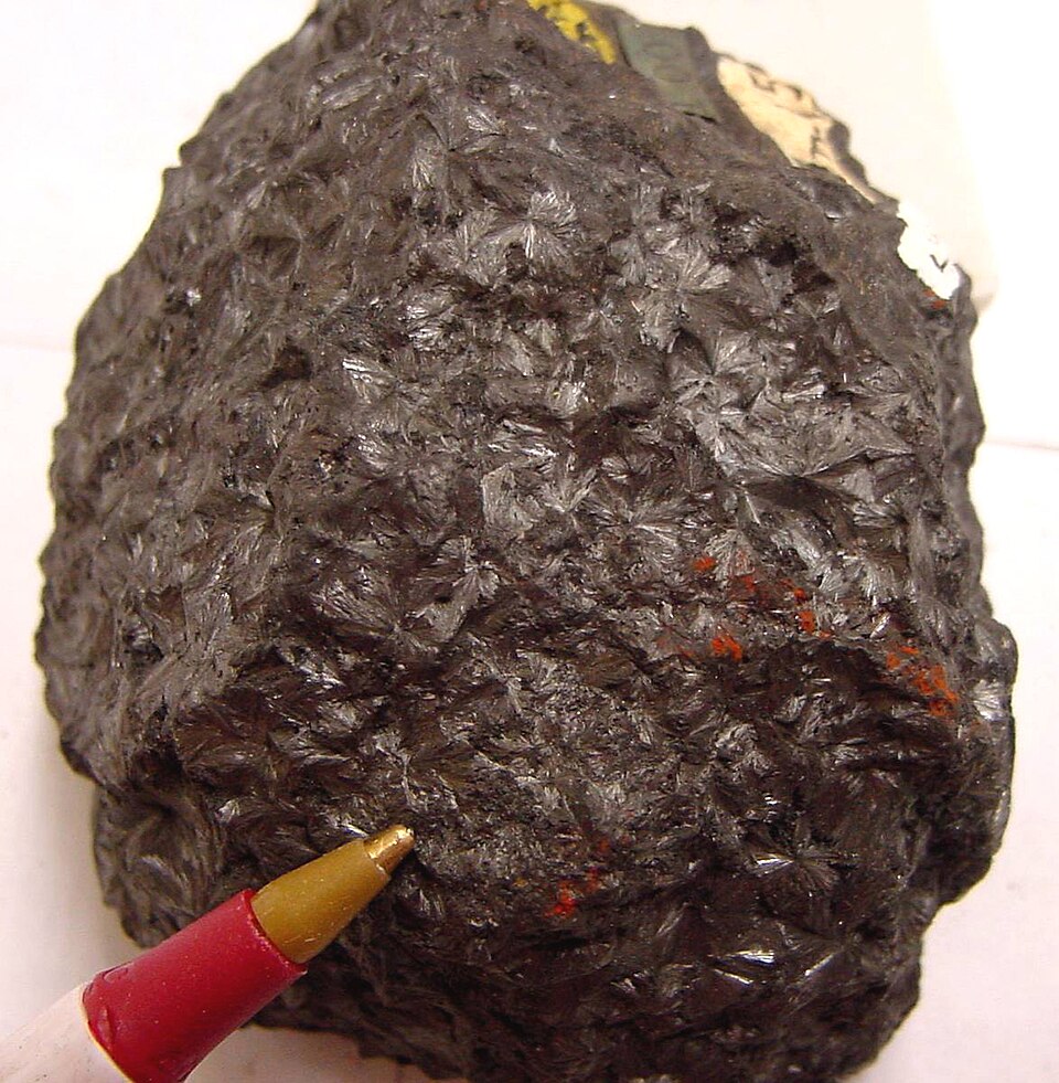 A dark steel-grey specimen of pyrolusite ore showing radiating metallic crystal clusters, with a pencil tip beside it for scale
