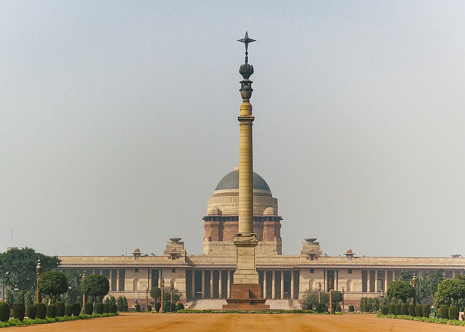 The domed sandstone facade of Rashtrapati Bhavan in New Delhi seen from the forecourt, with the Jaipur Column and its star finial standing in front