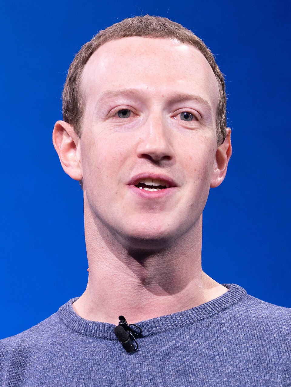 Mark Zuckerberg speaking on stage in a grey crew-neck sweater with a clip-on microphone, against a blue backdrop
