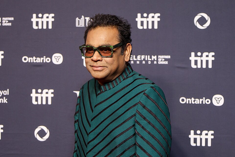 Composer A. R. Rahman in dark glasses and a green striped jacket standing in front of a film-festival backdrop