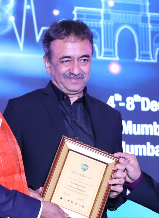 Film director Rajkumar Hirani in a dark jacket receiving a framed certificate on stage