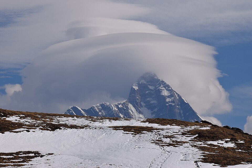 The snow-streaked pyramidal summit of Nanda Devi rising above a grassy alpine ridge, crowned by a large lenticular cloud