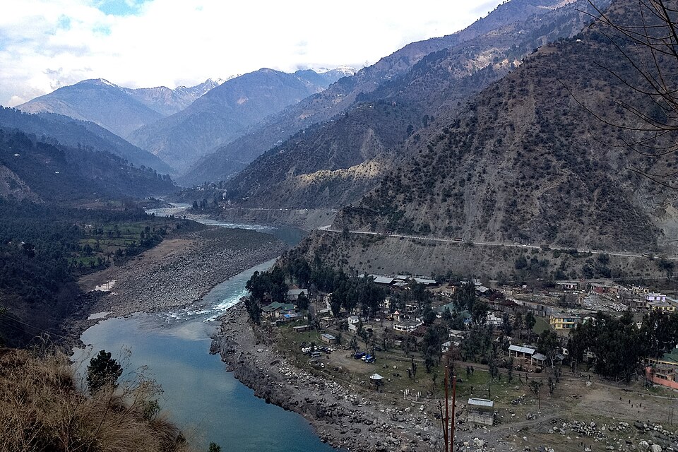 The Chenab river winding through a steep Himalayan valley at Ramban, with a hillside settlement on its right bank and snow-dusted ridges beyond