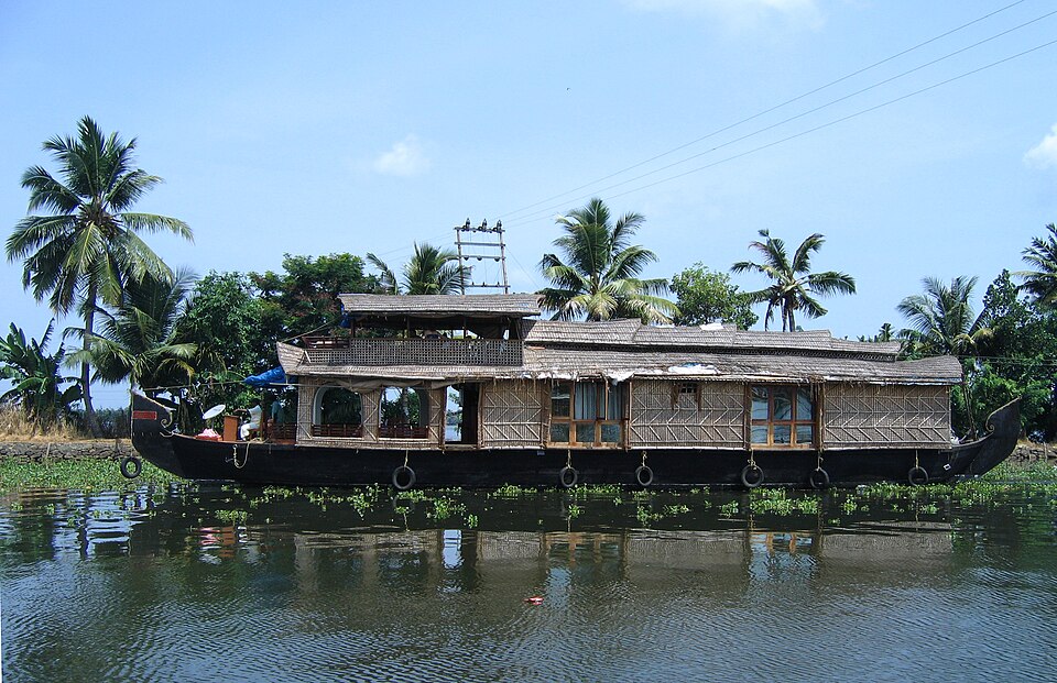 A thatched Kerala houseboat moored on the still, weed-fringed water of the Vembanad backwaters, with coconut palms lining the far bank