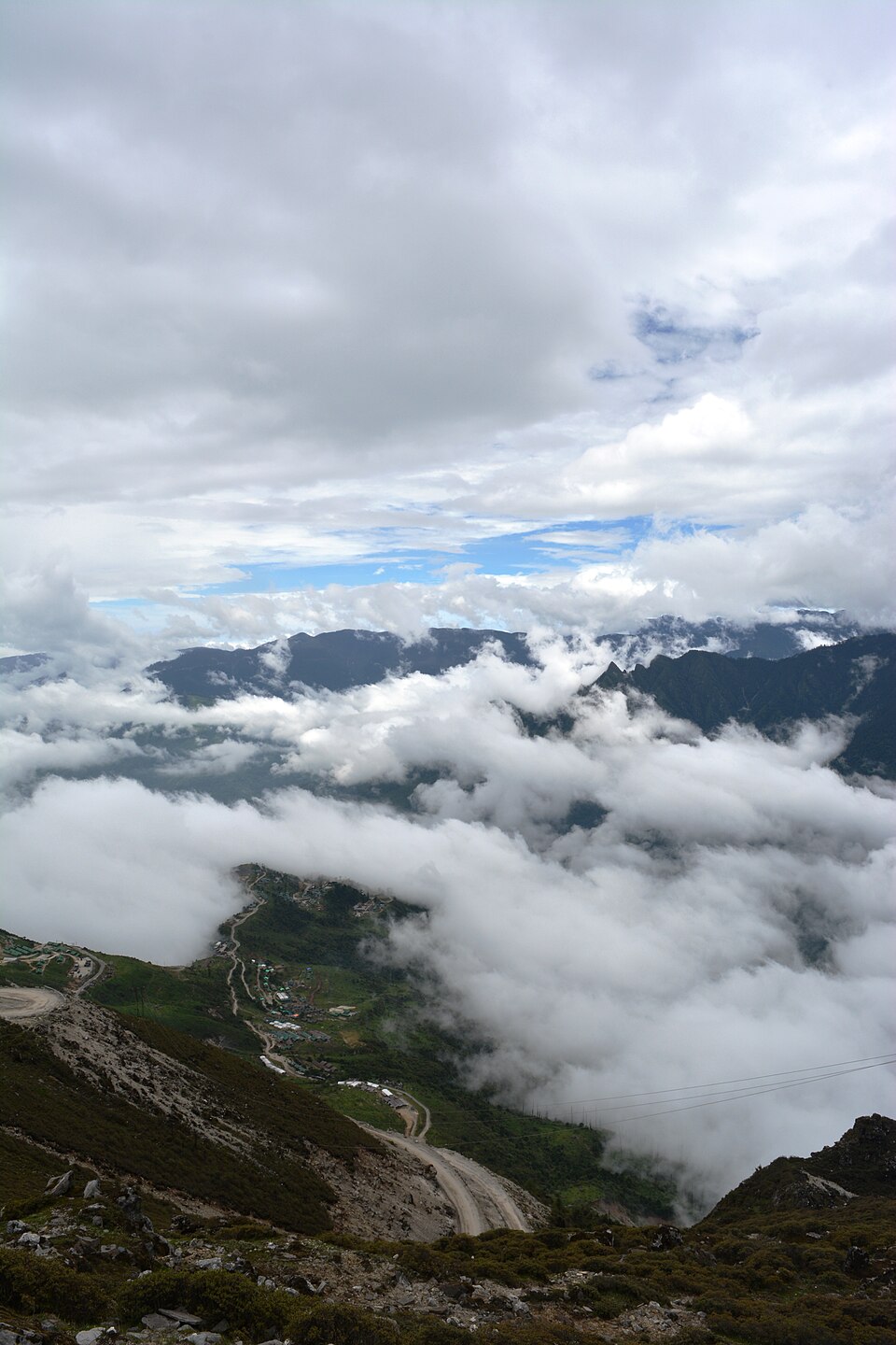 View from Sela Pass in Tawang district, Arunachal Pradesh: cloud filling a deep valley below forested ridges, with a small settlement and a dirt road clinging to the slope