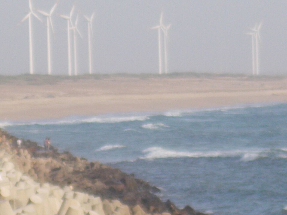 A row of white wind turbines standing on the low dunes behind a Gujarat beach, with surf breaking over a rocky breakwater in the foreground