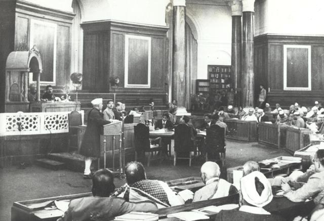 Black-and-white photograph of the Constituent Assembly of India in session, a member speaking from a lectern in the wood-panelled chamber while other members sit at desks around him