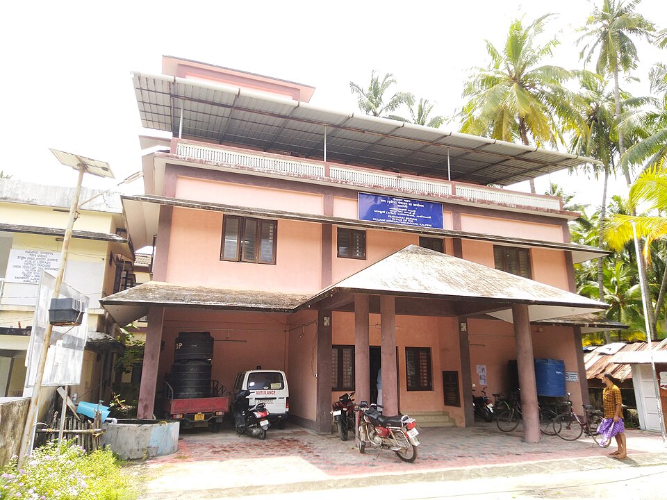 A two-storey village panchayat office building with a porch, signboard and parked bicycles and motorcycles, coconut palms rising behind it