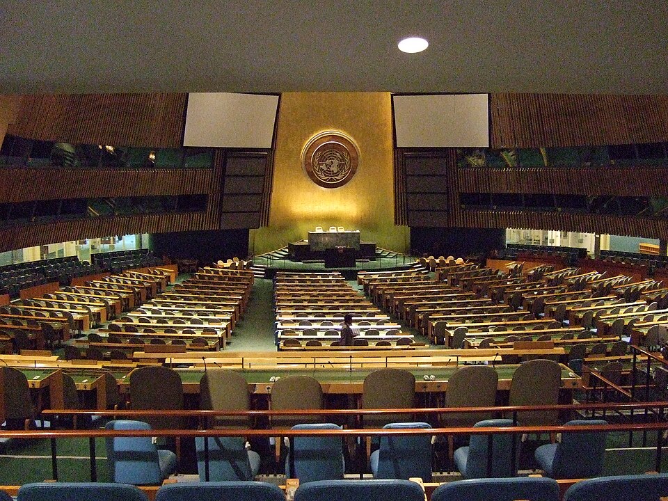 The empty United Nations General Assembly Hall in New York, rows of delegate desks facing the podium beneath the gold UN emblem on the end wall